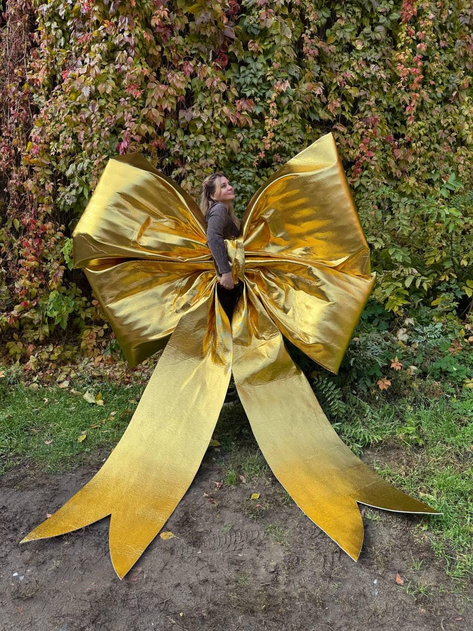 May include: A large, golden bow with long, flowing tails. The bow is made of a shiny, metallic material and is positioned in front of a backdrop of green and red foliage. The bow's size suggests it could be used for a festive decoration.