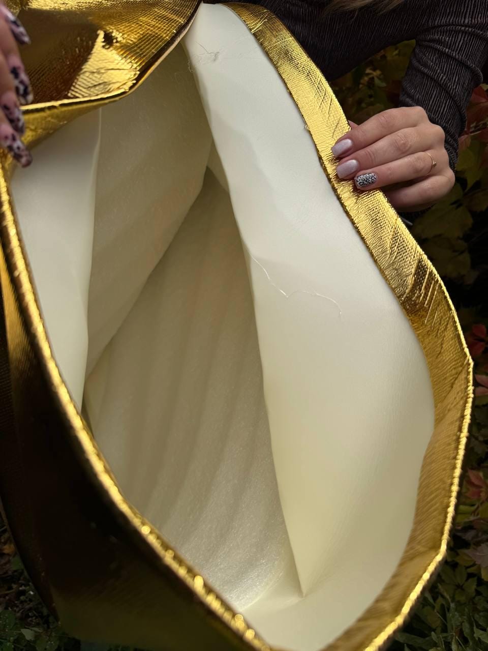 May include: A gold-colored, textured tote bag with a white interior. The bag has a rectangular shape with a gold trim around the edges. The interior is a light cream color.