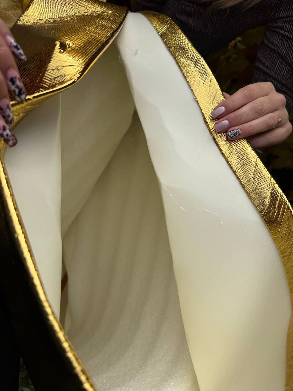 May include: Close-up of a gold metallic fabric with a textured surface, framing a white interior. The gold material has a stitched border. The image shows a glimpse of the inside of a garment or accessory.