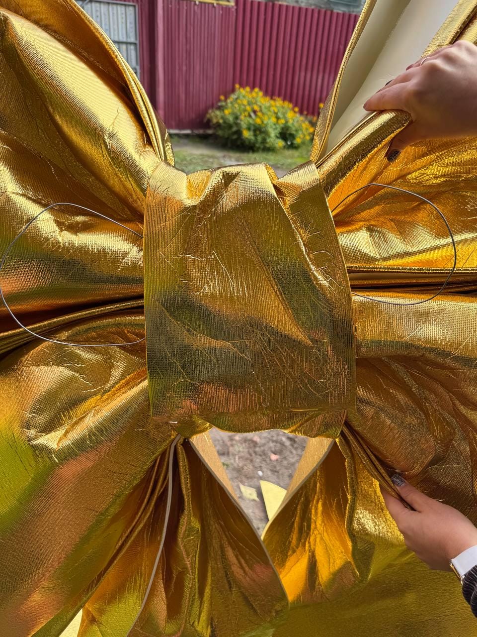 May include: A large, golden bow made of shiny, textured fabric. The bow is intricately folded and tied, with a wire loop visible. The bow is held by hands, suggesting it is a decorative element, possibly for a gift or event.