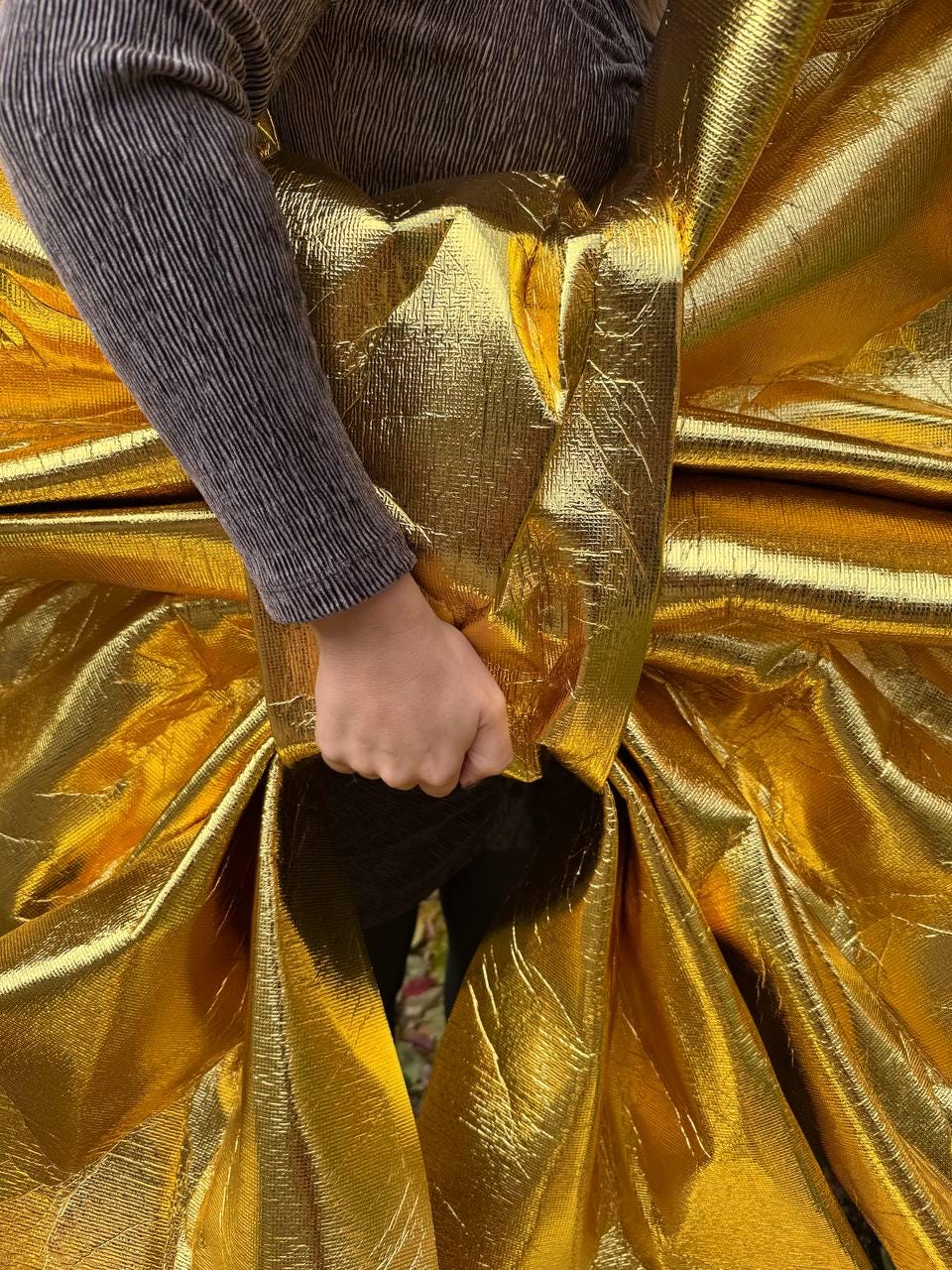 May include: A close-up of a shiny, golden fabric with a textured surface. The fabric is gathered and draped, creating folds and shadows. A person's arm in a gray ribbed sleeve is visible, holding the fabric.