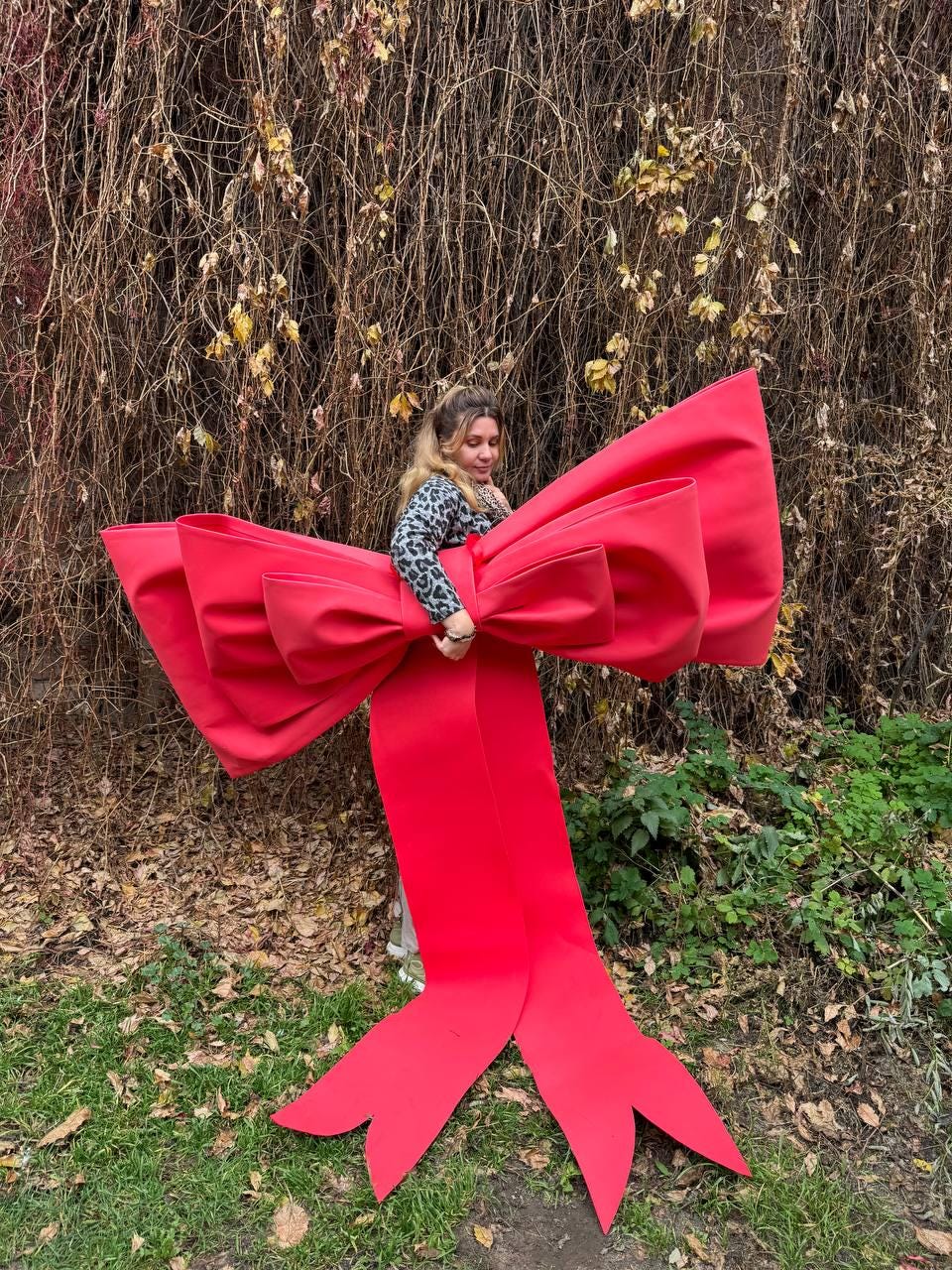 May include: A large, vibrant red bow prop with long, flowing ribbons. The bow is held by a person wearing a leopard print top, set against a backdrop of autumn foliage and greenery. The bow is a festive decoration.