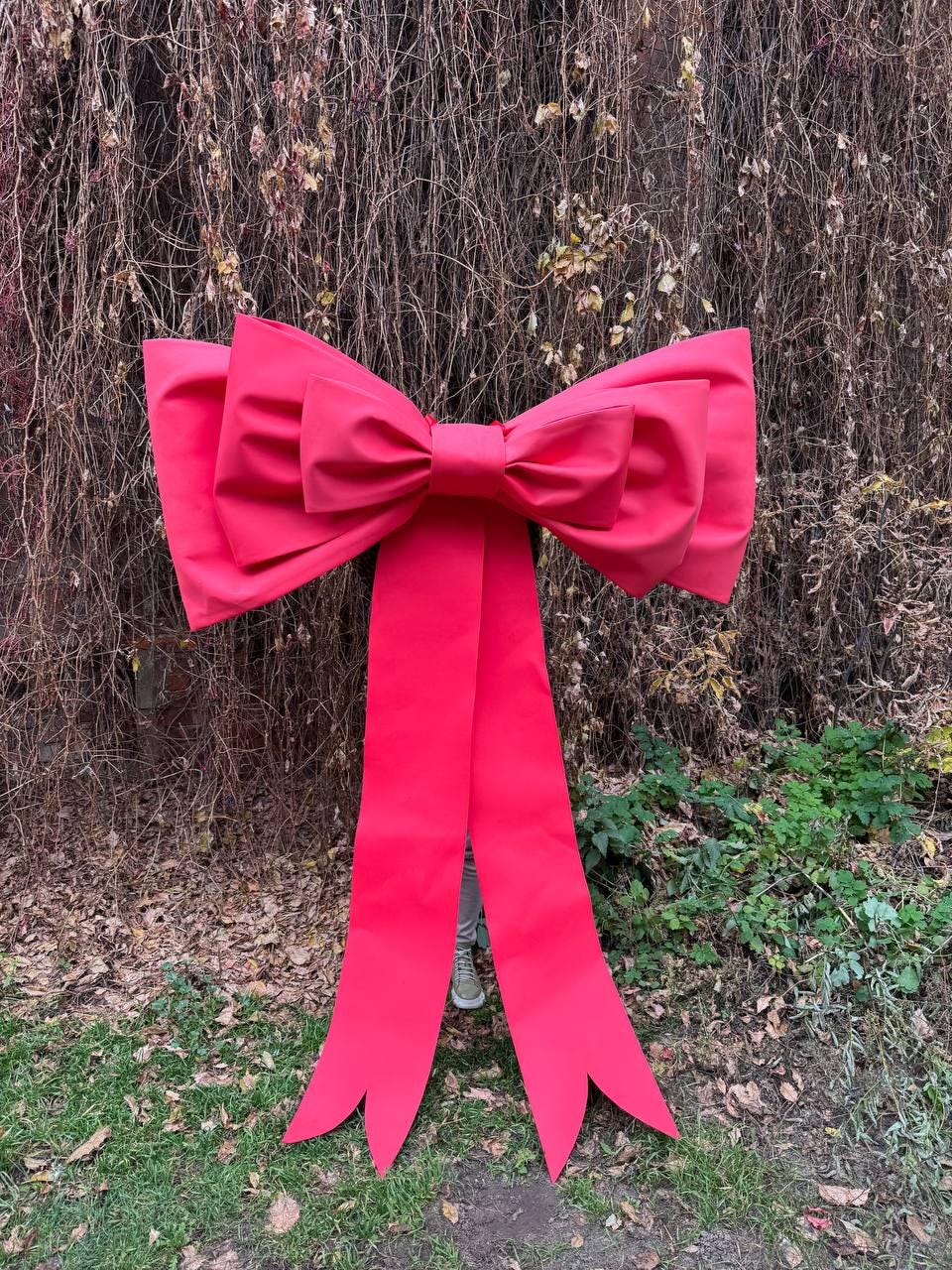 May include: A large, vibrant red bow with long, flowing tails. The bow is made of a smooth, possibly satin-like material, and is displayed outdoors against a backdrop of brown vines and green foliage. The bow's size suggests it's for decoration.