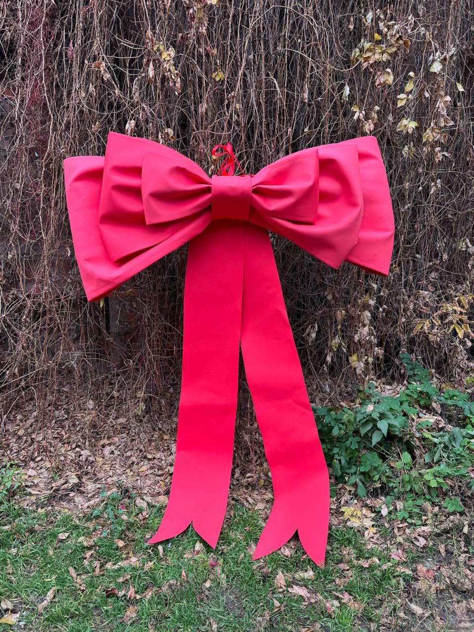 May include: A large, vibrant red bow with long, flowing tails. The bow is made of a smooth, possibly satin-like material and is designed for decoration. The bow is hanging against a backdrop of brown branches and green foliage.