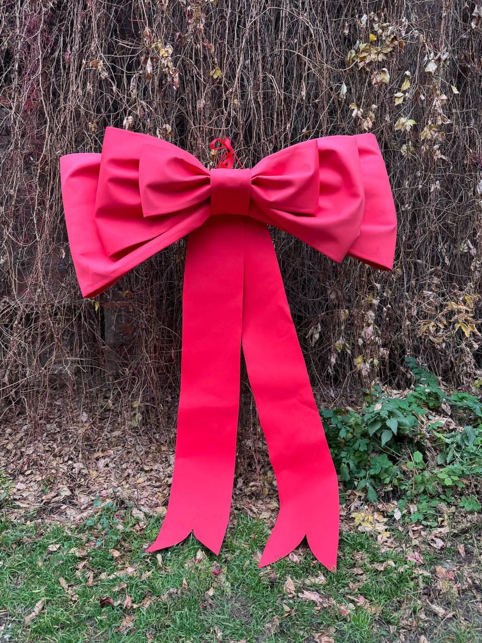 May include: A large, vibrant red bow with long, flowing tails. The bow is made of a smooth, possibly satin-like material and is suspended against a backdrop of dried vines and foliage. The tails have a decorative split at the bottom.