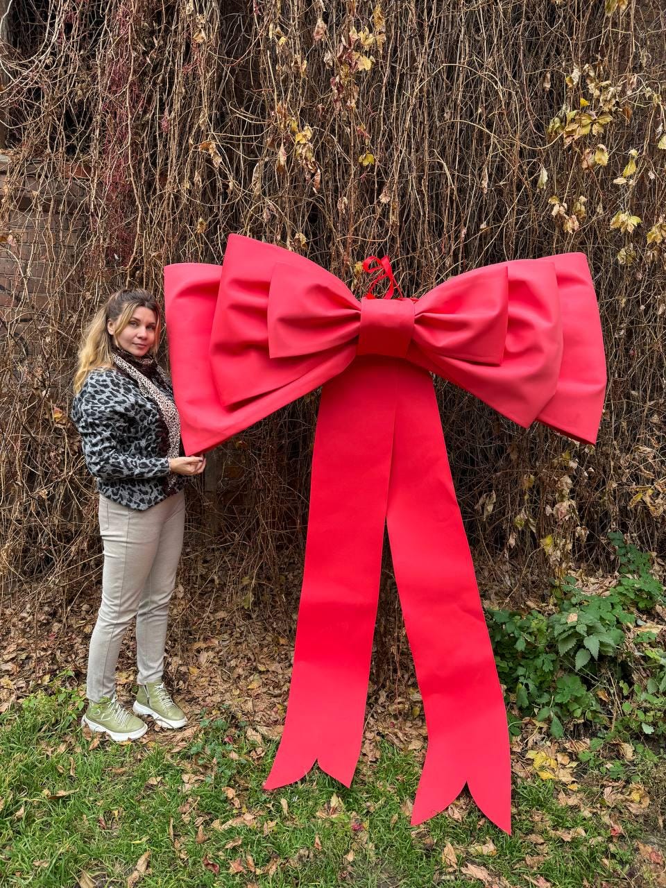 May include: A large, bright red decorative bow with long, flowing ribbons. The bow is displayed outdoors against a backdrop of greenery and vines. The bow is a festive decoration.