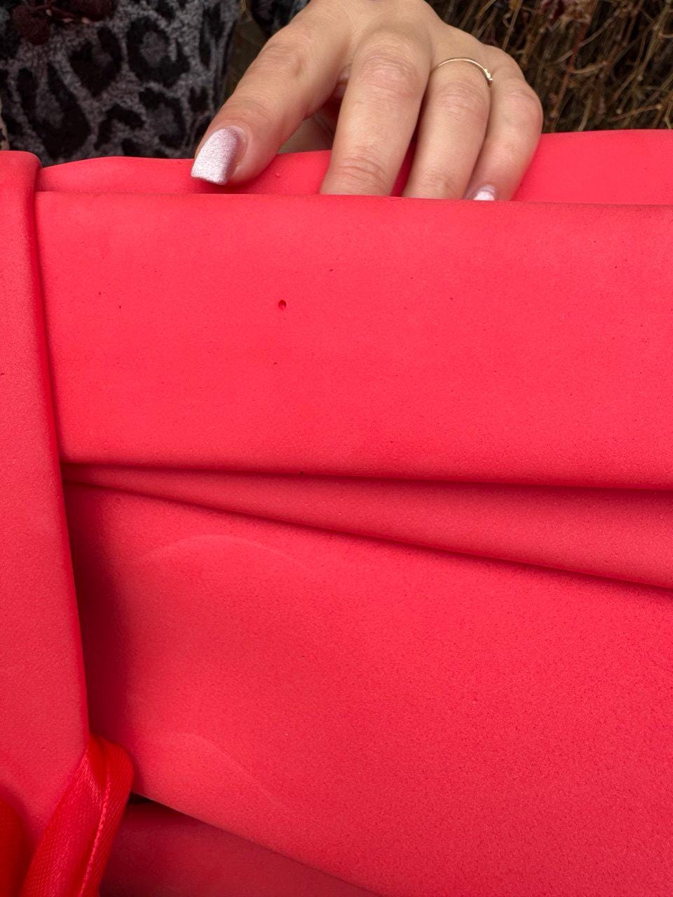 May include: Close-up of a bright red fabric with visible folds and a smooth texture. The fabric is held by a hand with a gold ring and manicured nails. The overall impression is of a vibrant, solid-colored material.