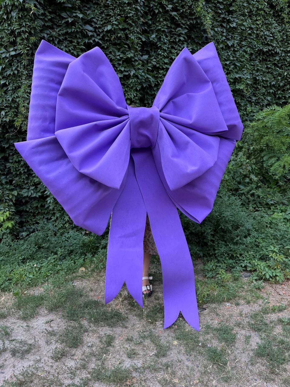 May include: A large, vibrant purple bow with long, flowing tails. The bow is made of a smooth fabric and is set against a backdrop of green foliage. The bow's size suggests it could be used for decoration.