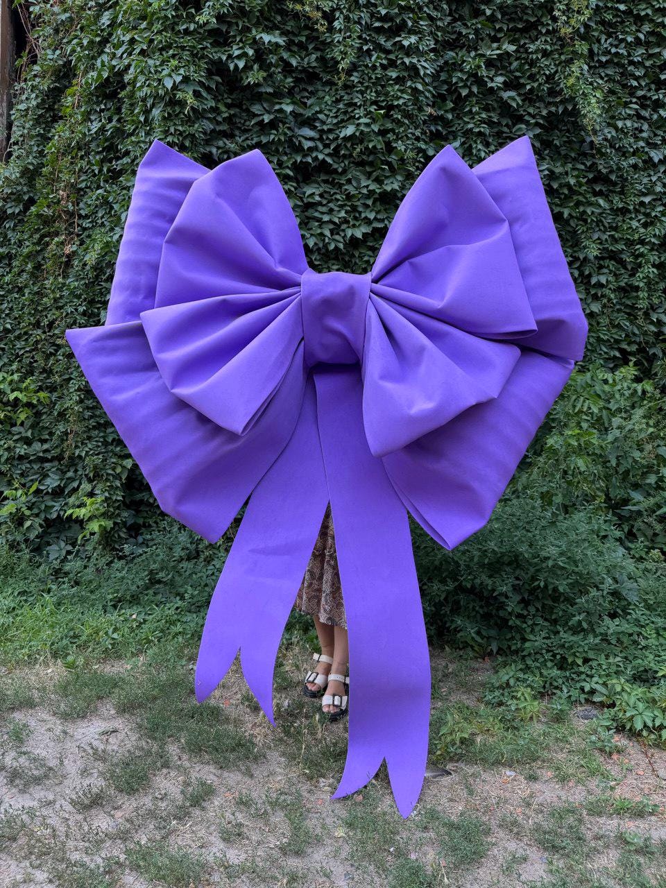 May include: A large, vibrant purple bow with long, flowing tails. The bow is made of a smooth, possibly satin-like fabric and is set against a backdrop of green foliage. The bow's size suggests it could be used for decoration or as a prop.