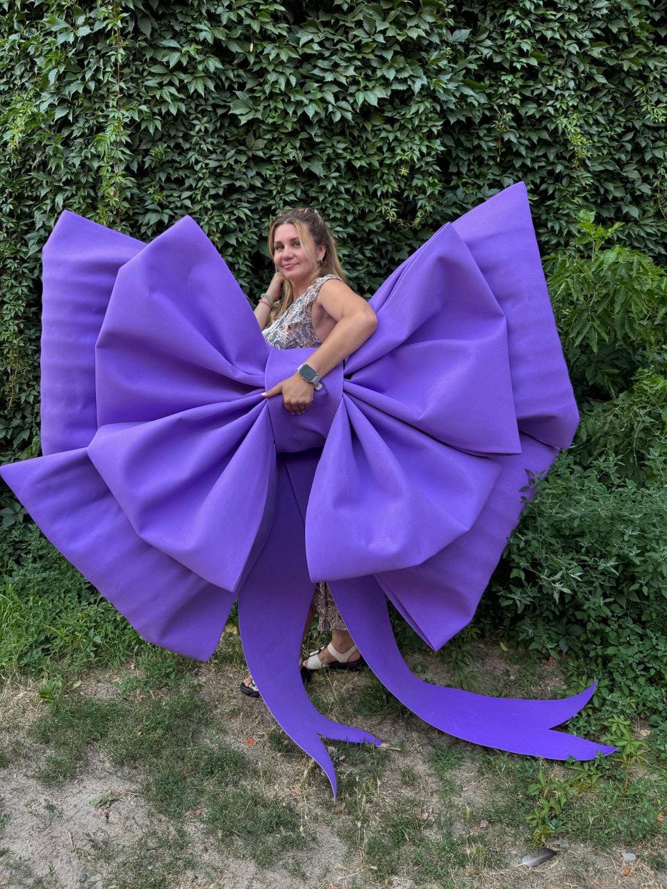 May include: A large, vibrant purple bow, with long flowing tails, is the focal point. The bow is held by a person. The bow's color is a striking shade of purple, and the material appears to be a sturdy fabric. The bow is a decorative item.