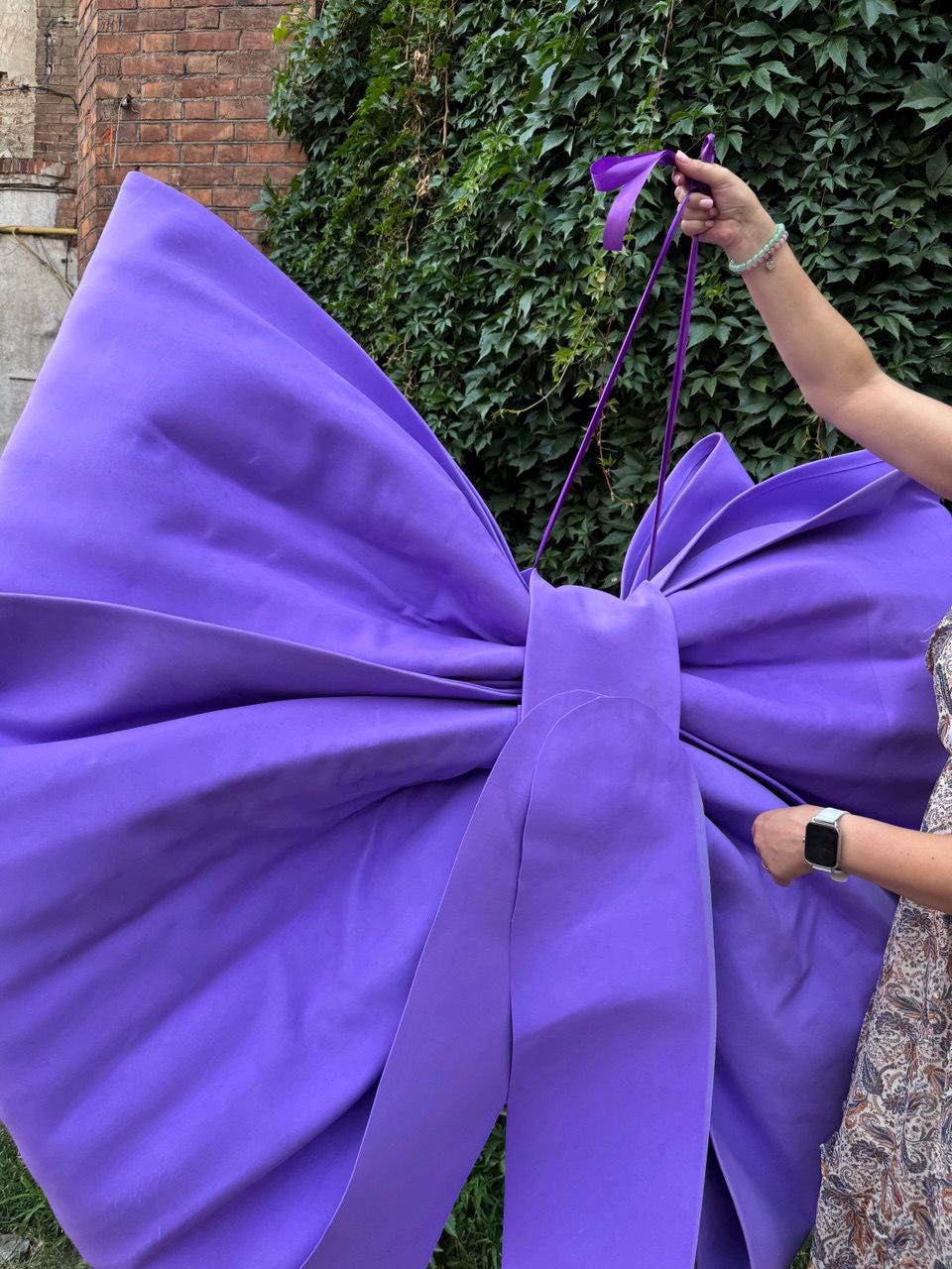 May include: A large, vibrant purple bow made of fabric. The bow is held up by a person's hand, with the bow's ribbons extending upwards. The bow is a striking decorative element, perfect for events or displays.