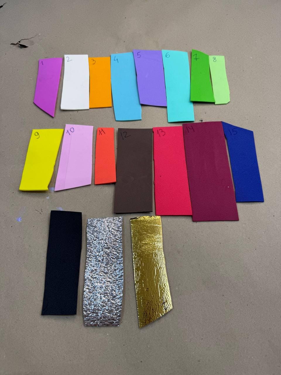 May include: A collection of fifteen rectangular swatches in various colors, including purple, white, orange, blue, and pink. Additional swatches are black, silver, and gold. The swatches are arranged on a neutral surface.