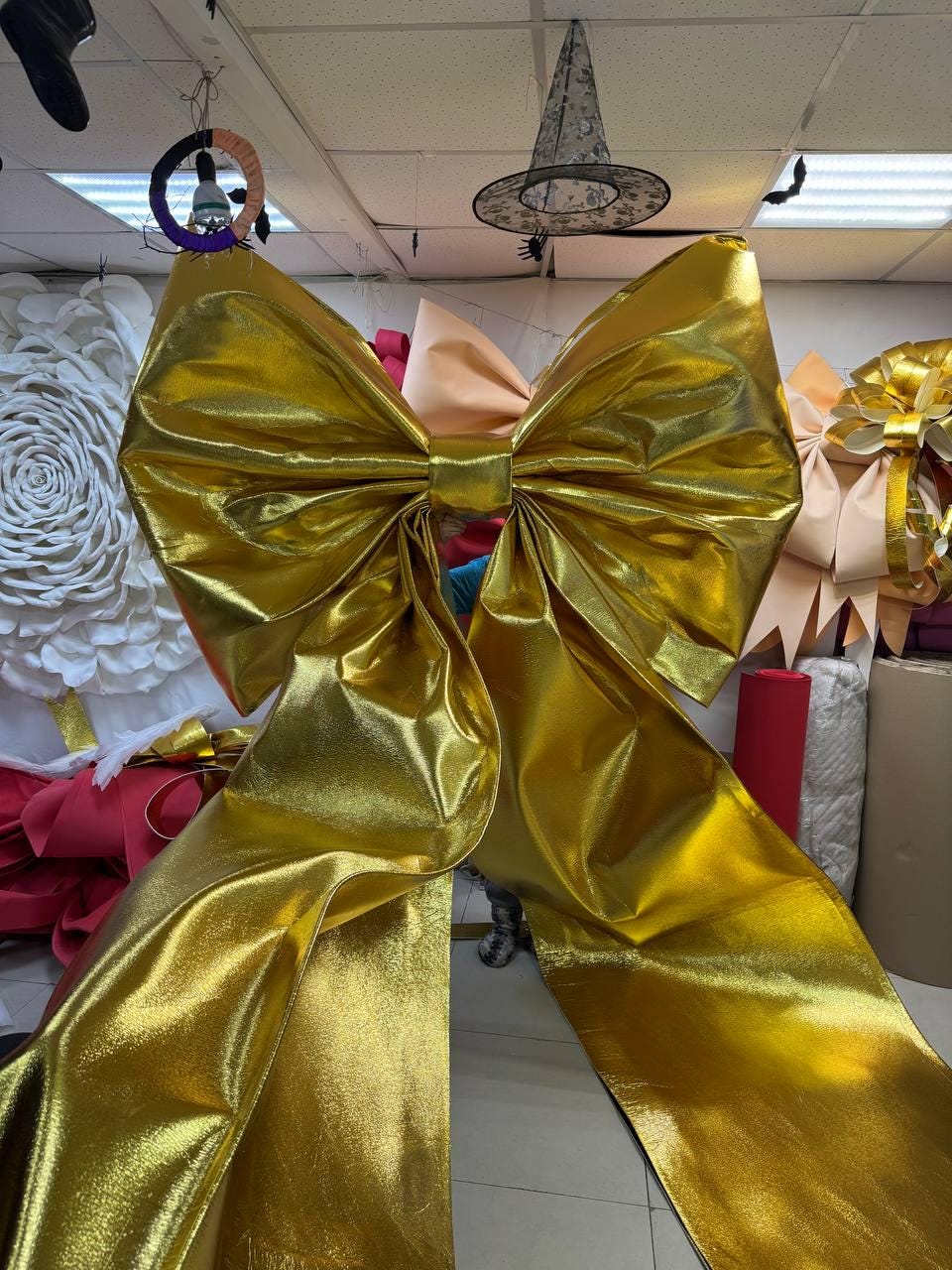 May include: A large, shiny gold bow made of a metallic fabric. The bow has multiple layers and long, flowing tails. Other bows and decorative items are visible in the background.