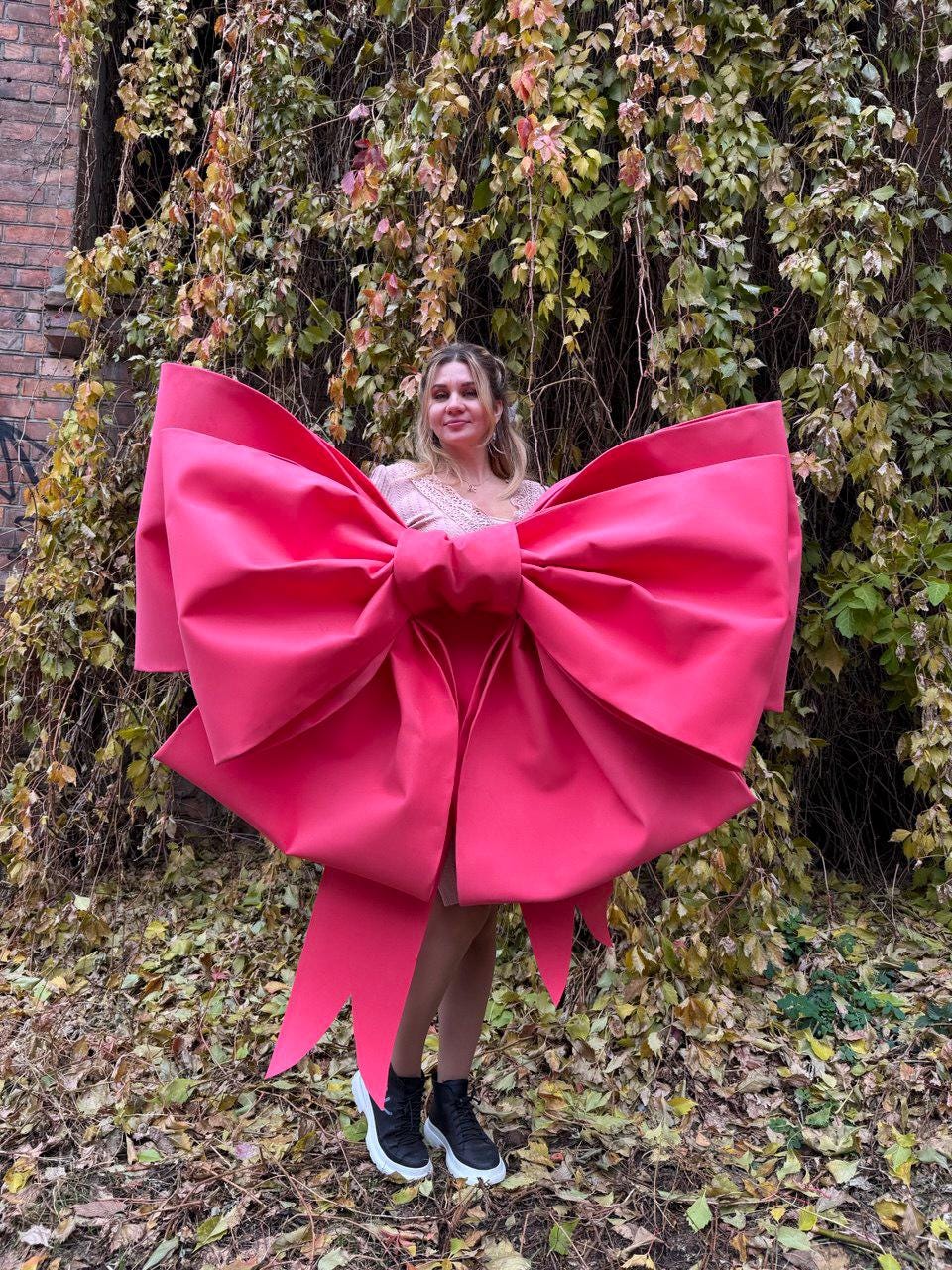 May include: A large, vibrant pink bow, held by a person, dominates the frame. The bow's size is striking, with long, flowing ribbons. The background features lush greenery and a brick wall, creating a contrast with the bow's bold color.
