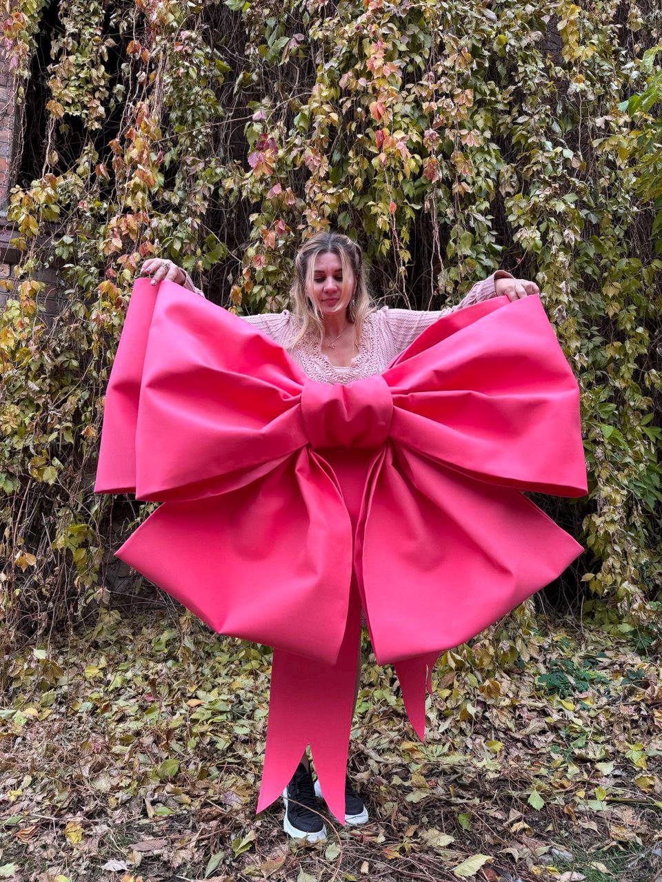 May include: A large, vibrant pink bow with long streamers. The bow is held up against a backdrop of autumn foliage. The bow's size and color make it a striking decorative element, suitable for events or displays.