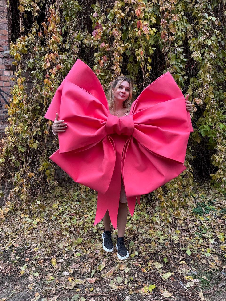 May include: A large, vibrant pink bow costume. The bow is the focal point, with a voluminous design and long, flowing tails. The person is wearing black sneakers with white soles. The background features autumn foliage.