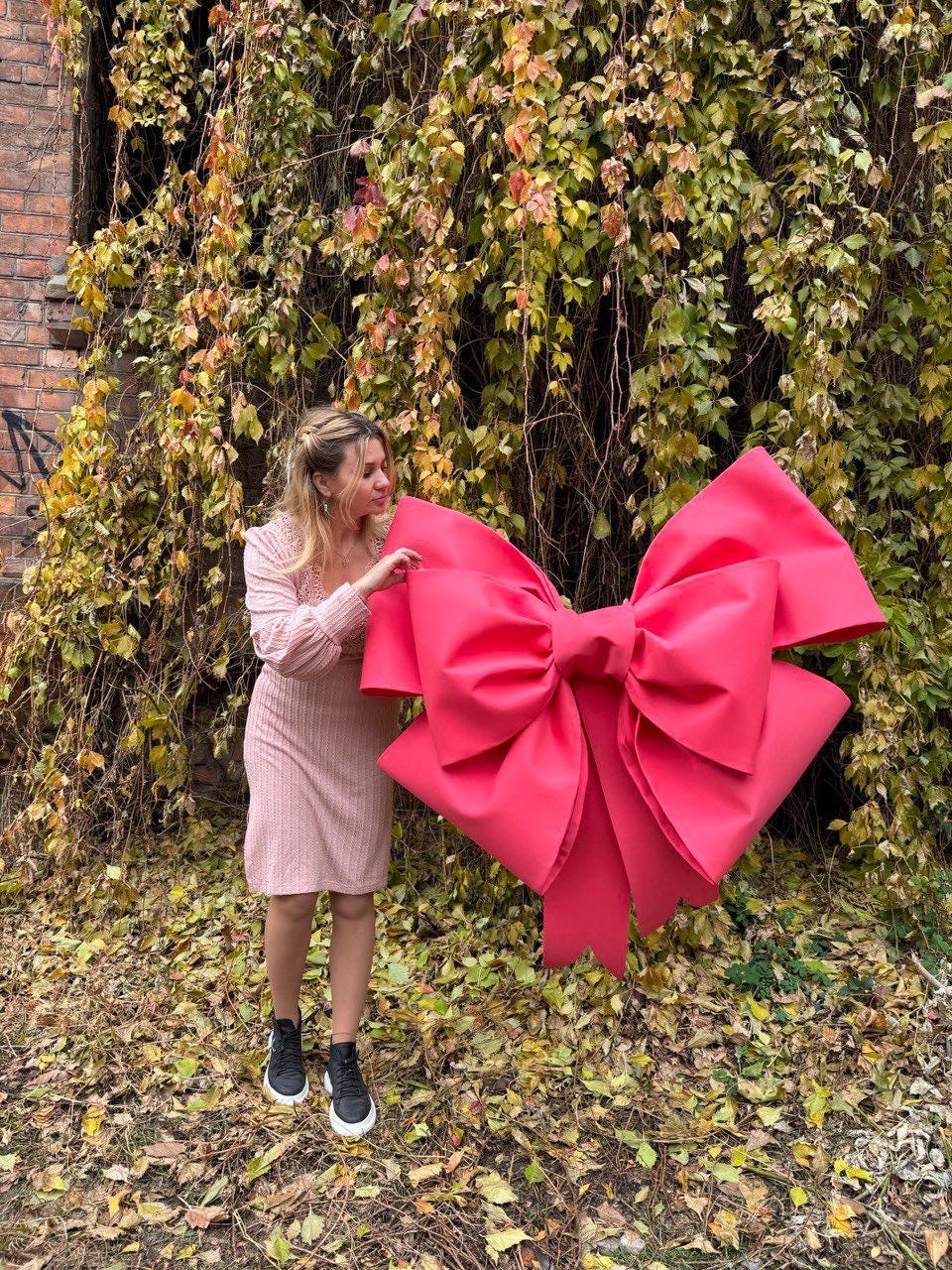 May include: A large, vibrant pink bow, possibly for decoration or a gift. The bow is held by a person wearing a pink dress and black sneakers. The background features autumn foliage and a brick wall.