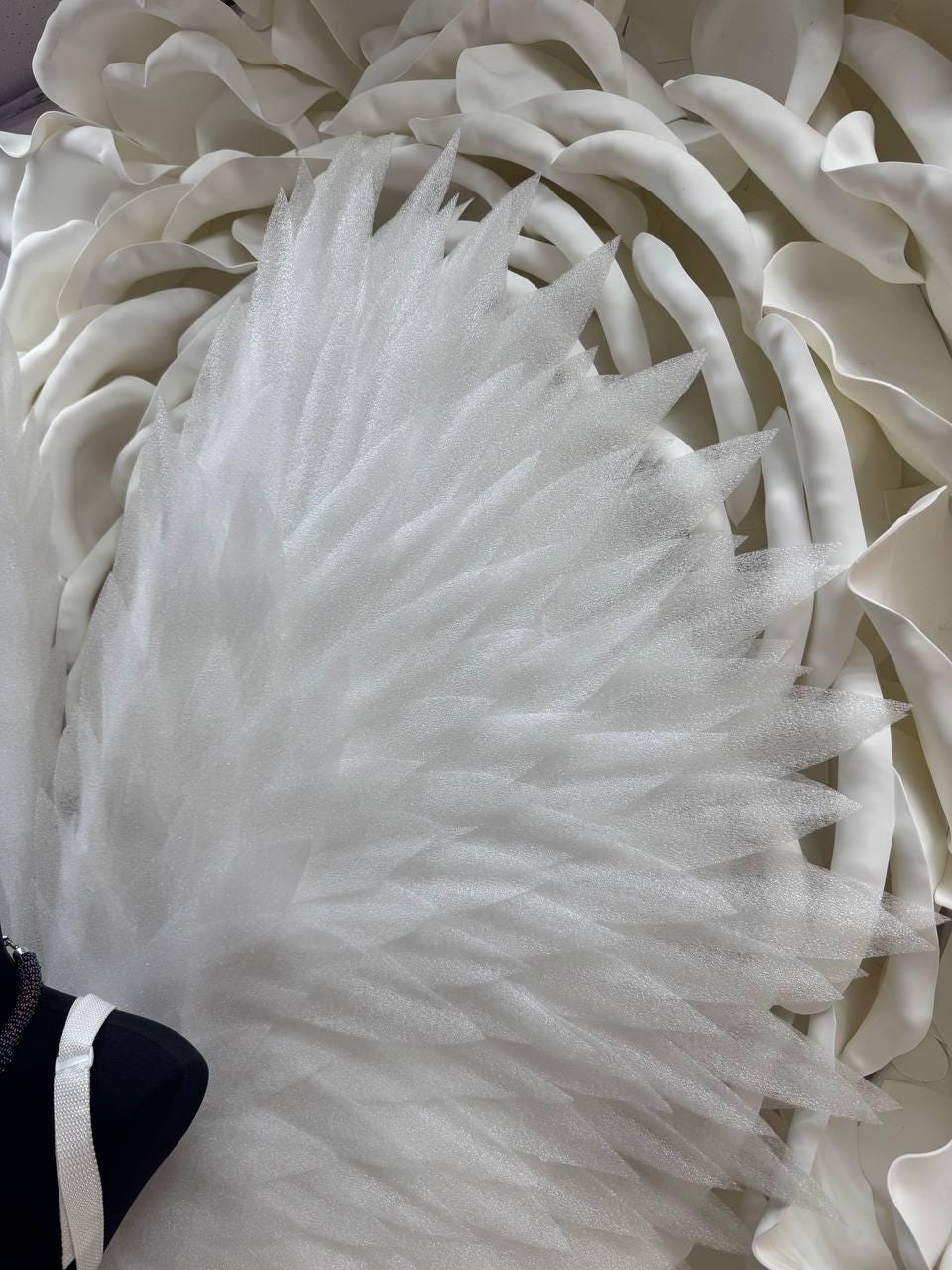 May include: Close-up of a white, feathery wing-like structure with sharp, pointed edges. The wing is made of a translucent, textured material, possibly fabric or foam. The background features large, white, petal-like shapes.