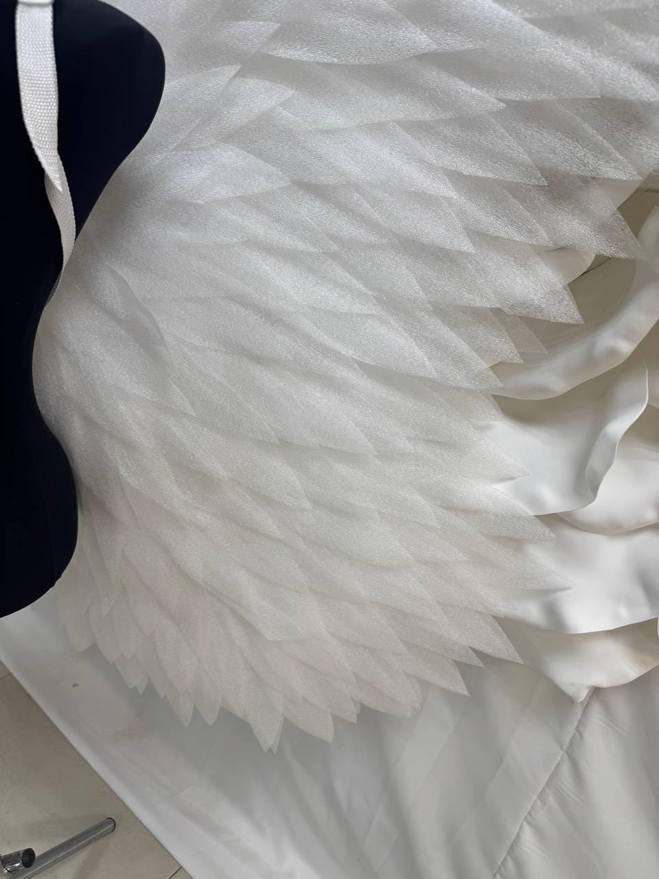 May include: White angel wings with layered, feather-like details. The wings are attached to a black mannequin torso with a white strap. The wings are made of a soft, textured material and are partially visible against a white background.