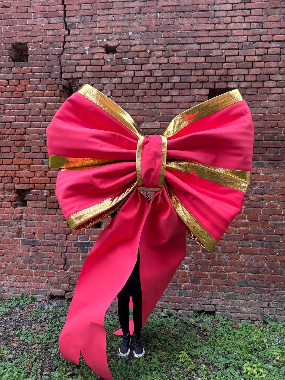 May include: A large, vibrant pink bow with gold trim. The bow is oversized, with long, flowing tails. The background is a weathered brick wall, and the bow is the focal point of the image. The bow is a festive decoration.