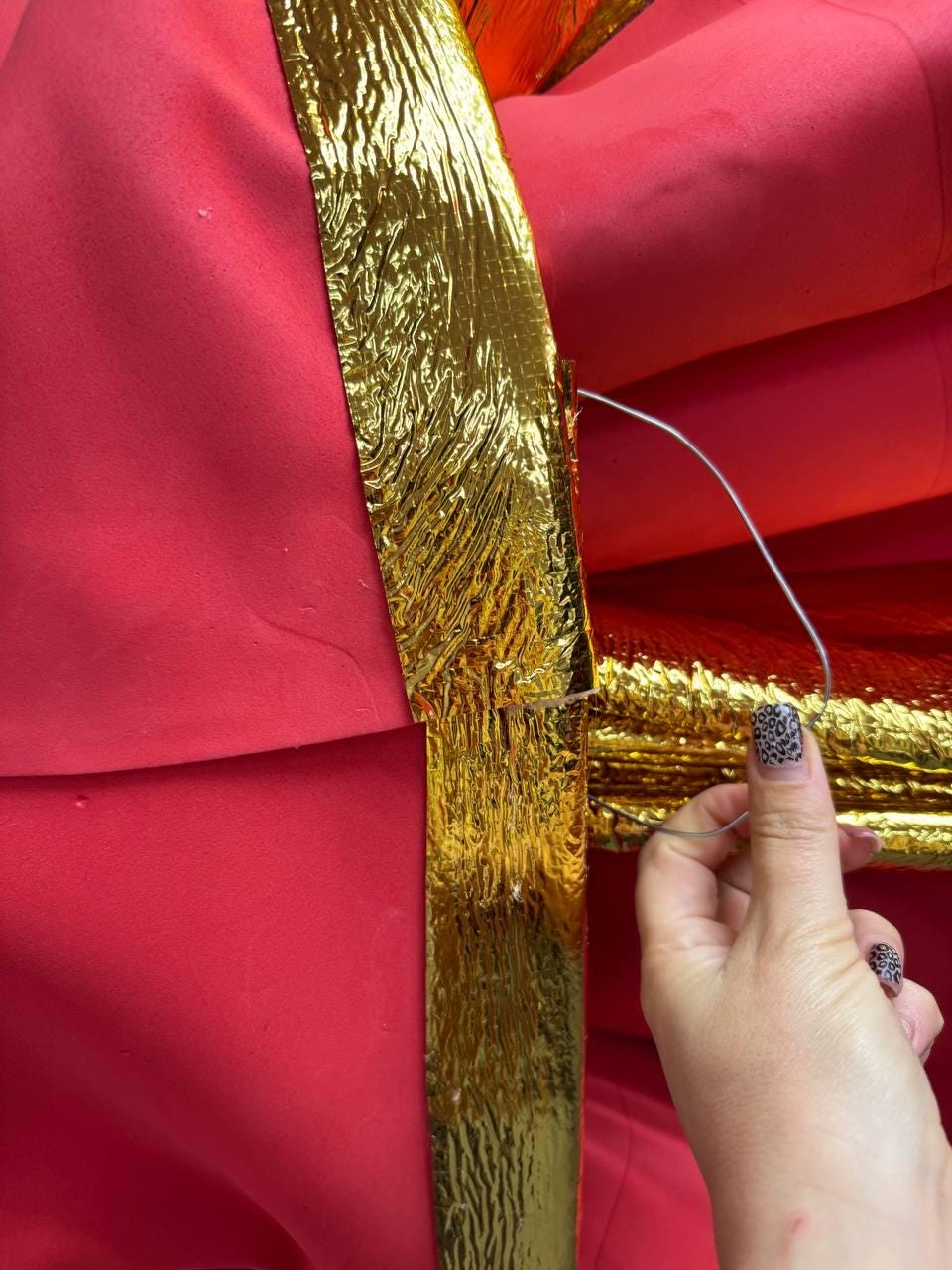 May include: Close-up of a red fabric with a gold metallic ribbon. The ribbon is textured and has a crinkled appearance. A hand with a silver wire is visible, holding the ribbon in place. The fabric appears to be part of a larger decorative item.