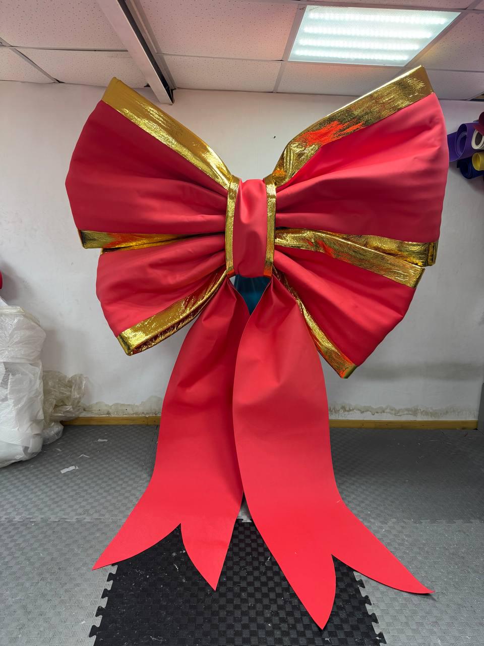 May include: A large, vibrant red bow with gold trim. The bow features a central knot and long, flowing tails. The edges of the bow are trimmed with gold. This decorative bow is suitable for holiday or event decorations.