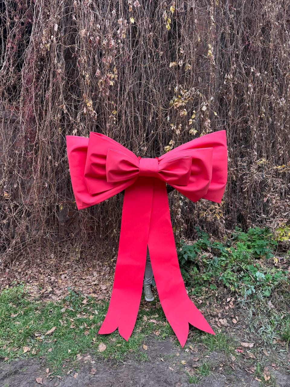 May include: A large, vibrant red bow with long, flowing tails. The bow is made of a smooth fabric and is positioned outdoors against a backdrop of brown vines and green foliage. The bow's size suggests it is intended for decorative purposes.