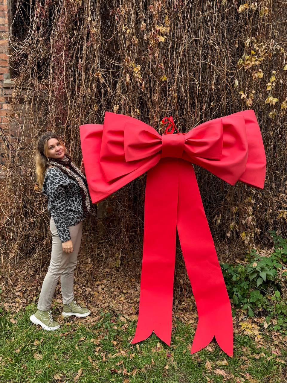 May include: A large, vibrant red bow with long, flowing ribbons. The bow is made of a smooth, possibly satin-like material. The bow is standing upright on a grassy area, suggesting it's a decorative item, possibly for a special event or holiday.