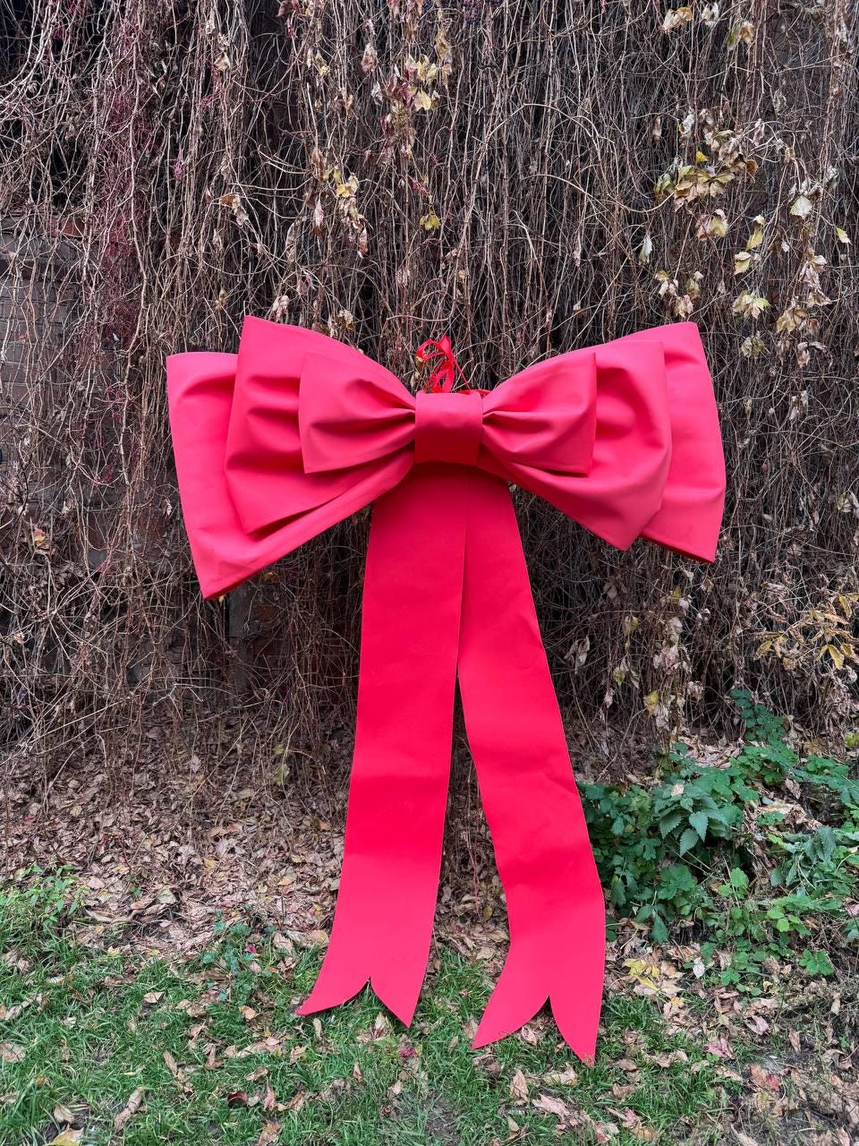 May include: A large, vibrant red bow with long, flowing tails. The bow is made of a smooth, possibly satin-like material. The bow is hanging against a backdrop of dried vines and green foliage, suggesting outdoor holiday decor.