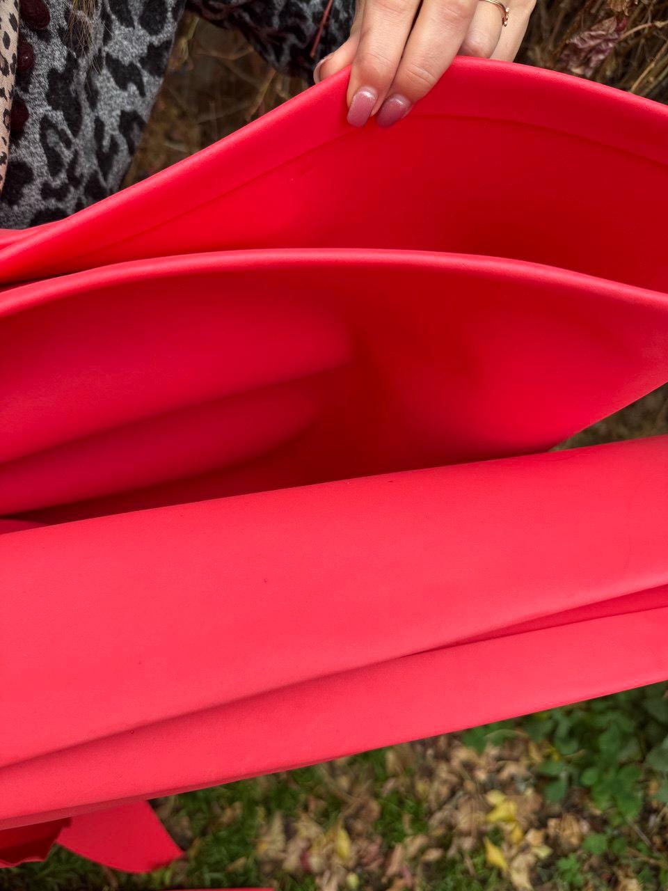May include: Bright red material, possibly fabric or a flexible sheet, is held up, revealing its folded layers. The material has a smooth, matte finish and a vibrant, saturated color. The image focuses on the texture and color of the material.