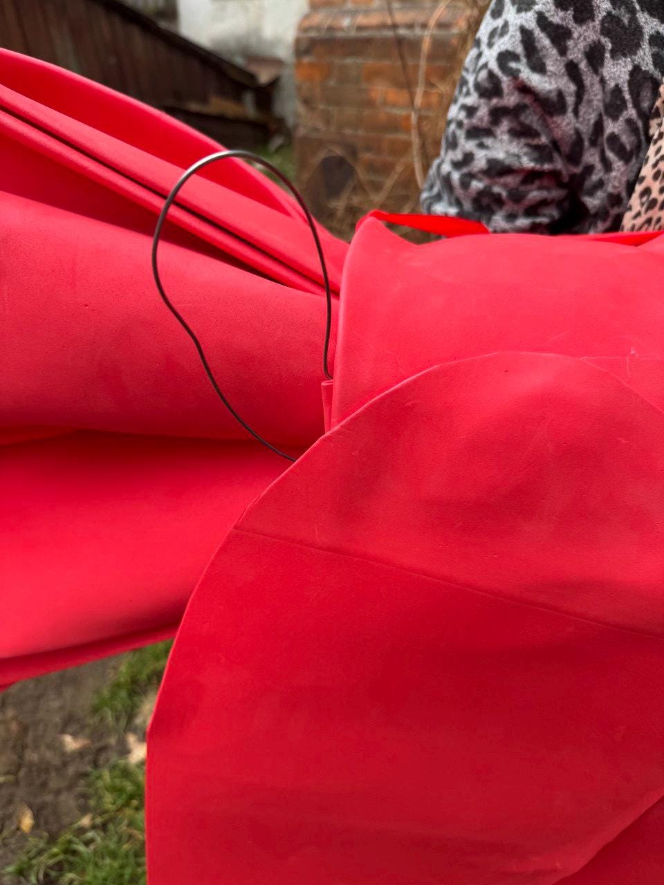 May include: Close-up of a large, bright red fabric bow with a wire loop. The bow is made of a soft, matte material and is folded into multiple layers. The wire loop is thin and dark, likely for hanging or attaching the bow.