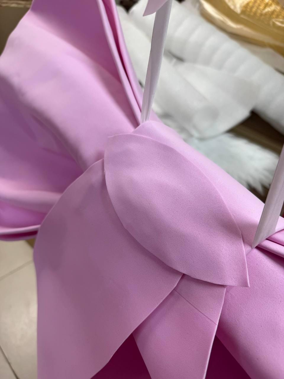 May include: Close-up of a large, light purple fabric bow with white ribbon. The bow is made of a soft, smooth material and has a layered design. The background is blurred, with hints of other materials.
