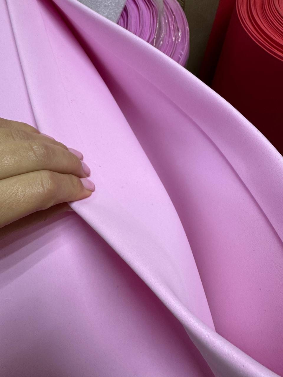 May include: Close-up of a hand holding a sheet of light pink foam material. The foam is smooth and appears to be flexible. Rolls of similar material in pink and red are visible in the background.