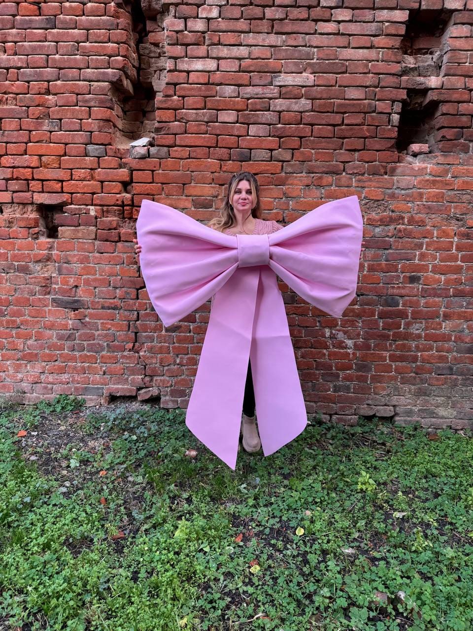 May include: A large, pink bow costume with long streamers. The bow is held in front of a person, against a backdrop of a weathered brick wall and green grass. The bow's color is a soft, pastel pink.