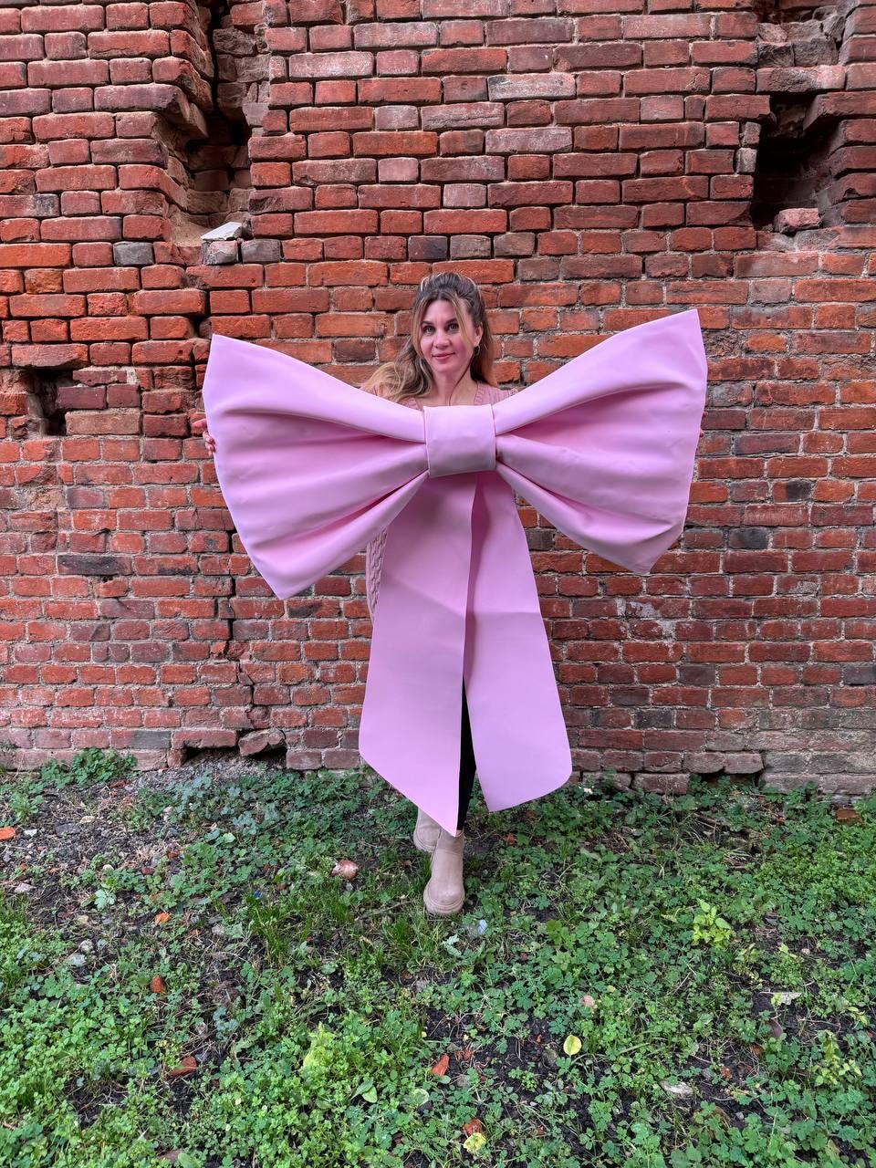 May include: A large, light pink bow costume with long, draping ribbons. The bow is held in front of a brick wall and green grass. The costume is a whimsical and eye-catching accessory, perfect for themed events or photo opportunities.