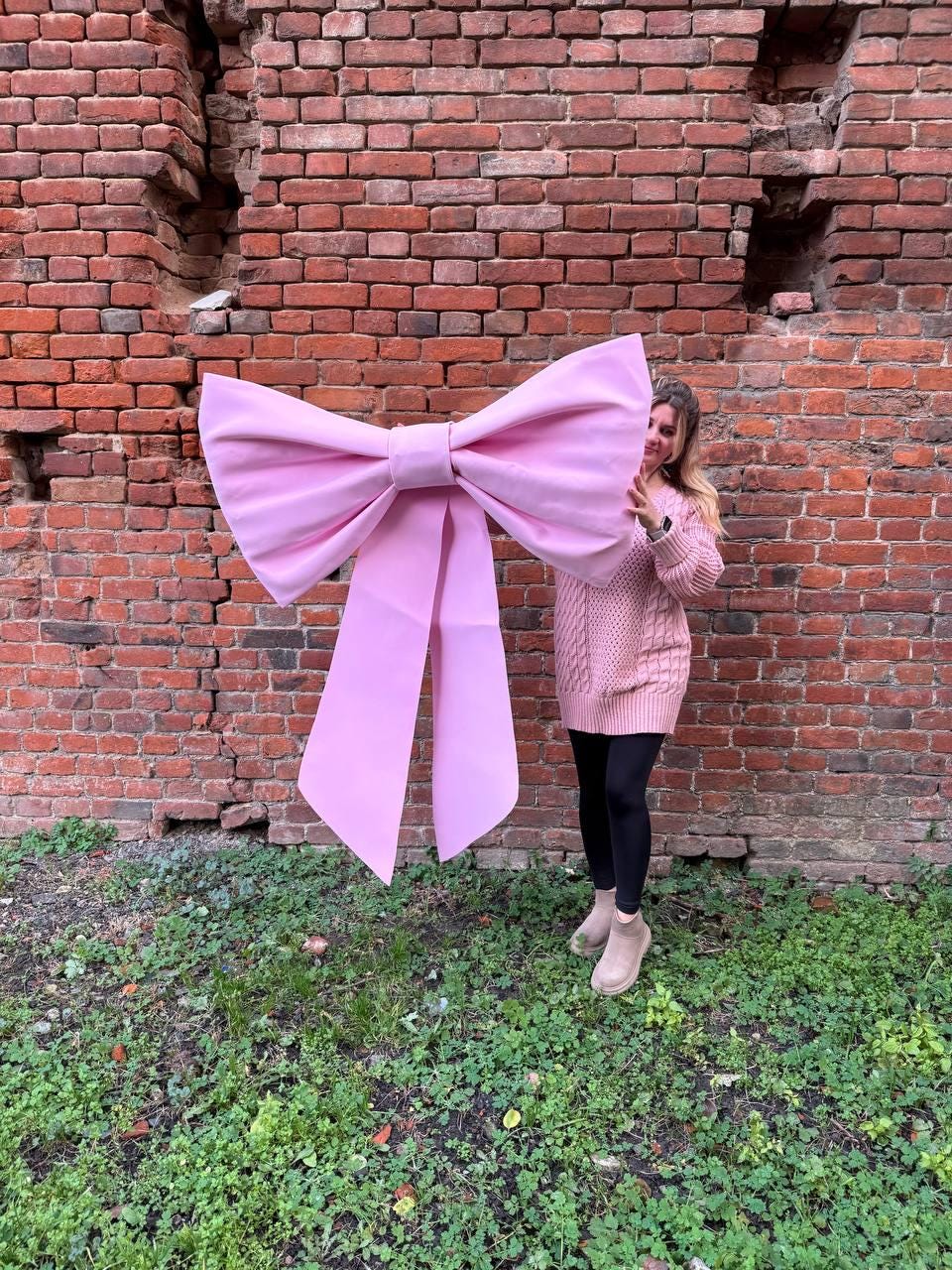 May include: A large, light pink bow with long streamers. The bow is held in front of a brick wall and green grass. The bow is a solid color and appears to be made of fabric.