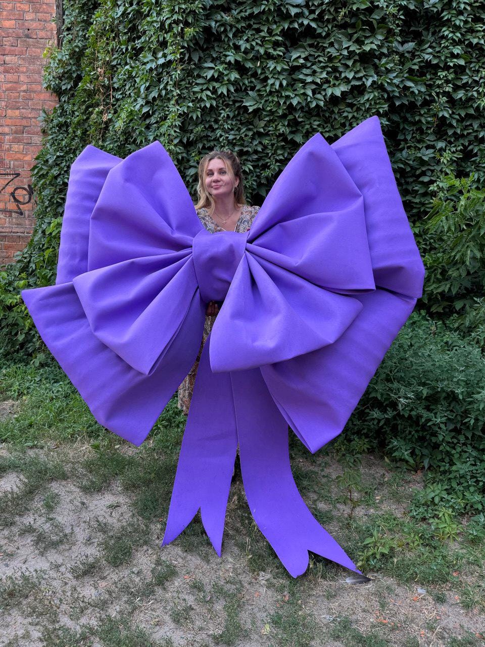 May include: A large, vibrant purple bow with long, flowing ribbons. The bow is made of a smooth fabric and is positioned in front of a background of greenery and a brick wall. The bow's size suggests it could be used for decoration.