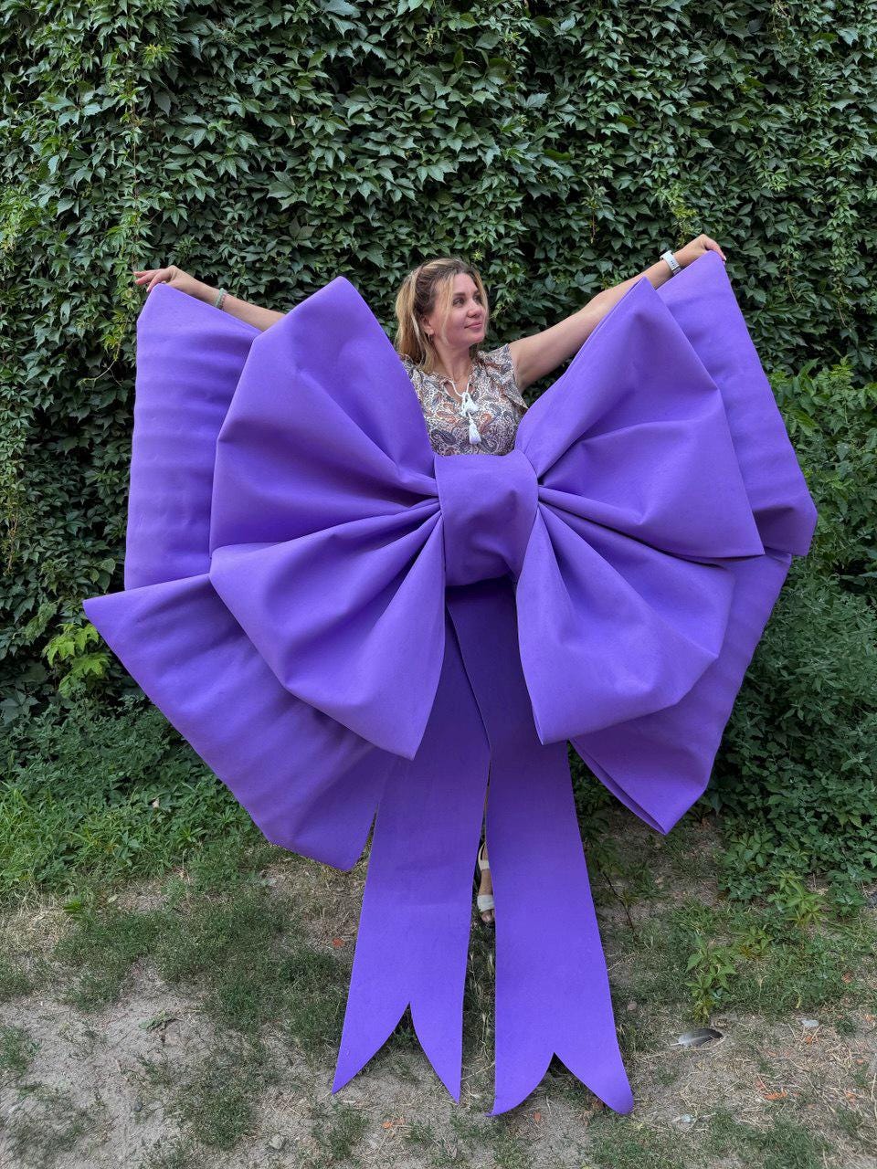 May include: A large, vibrant purple bow with long streamers. The bow is the focal point, with a person partially visible behind it. The background is a lush green wall of foliage. The bow's size suggests it's for decoration.