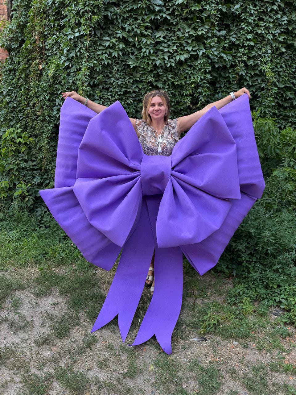 May include: A large, vibrant purple bow with long, flowing ribbons. The bow is held up by a person in front of a lush green backdrop. The bow's size and color make it a striking decorative element.