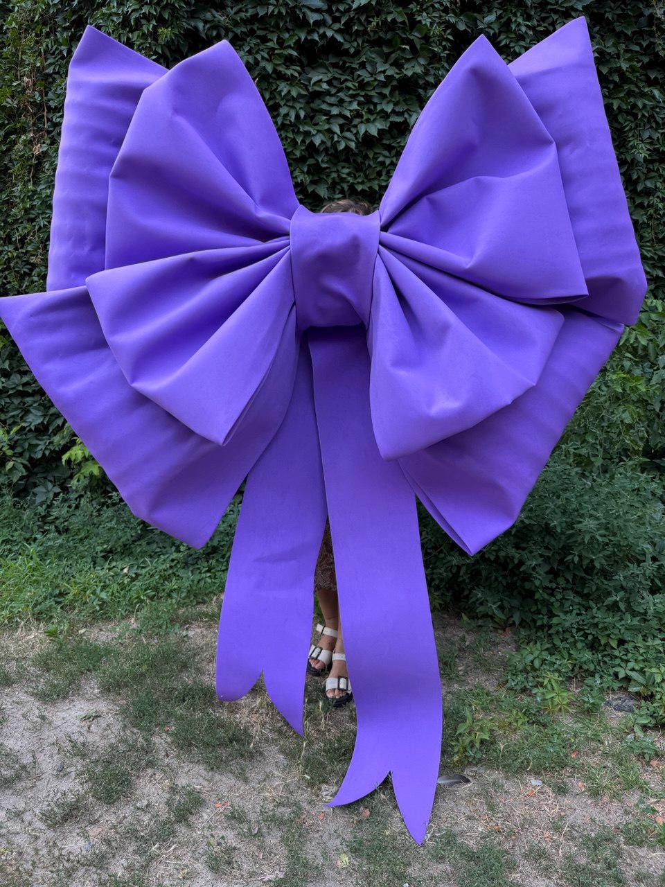 May include: A large, vibrant purple bow with long, flowing tails. The bow is made of a smooth, possibly satin-like material, and is positioned against a backdrop of green foliage. The bow's size suggests it could be used for decoration or as a prop.
