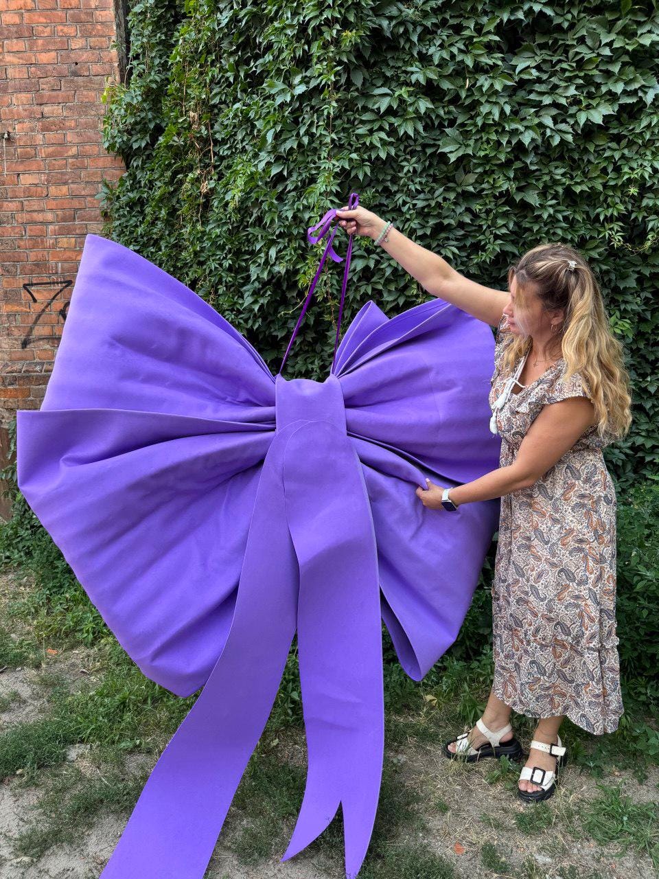 May include: A large, vibrant purple bow with long, flowing ribbons. The bow is held up by a person in a floral dress against a backdrop of green foliage and a brick wall. The bow is a striking decorative element.
