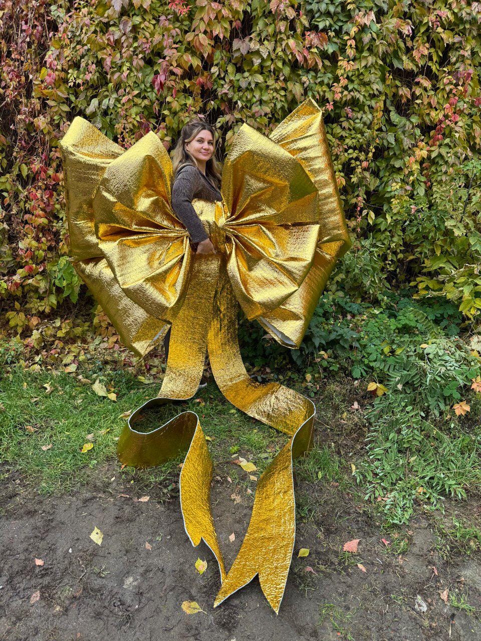 May include: A large, golden bow with long, flowing ribbons. The bow is made of a shiny, metallic material and is positioned in front of a background of green foliage and autumn leaves. The bow is large enough to be worn.