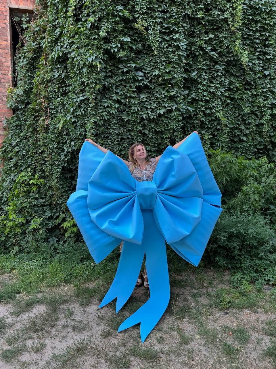 May include: A large, bright blue bow with long, flowing tails. The bow is made of a smooth, possibly satin-like material. The bow is held up in front of a person, with a background of green foliage and a brick building.
