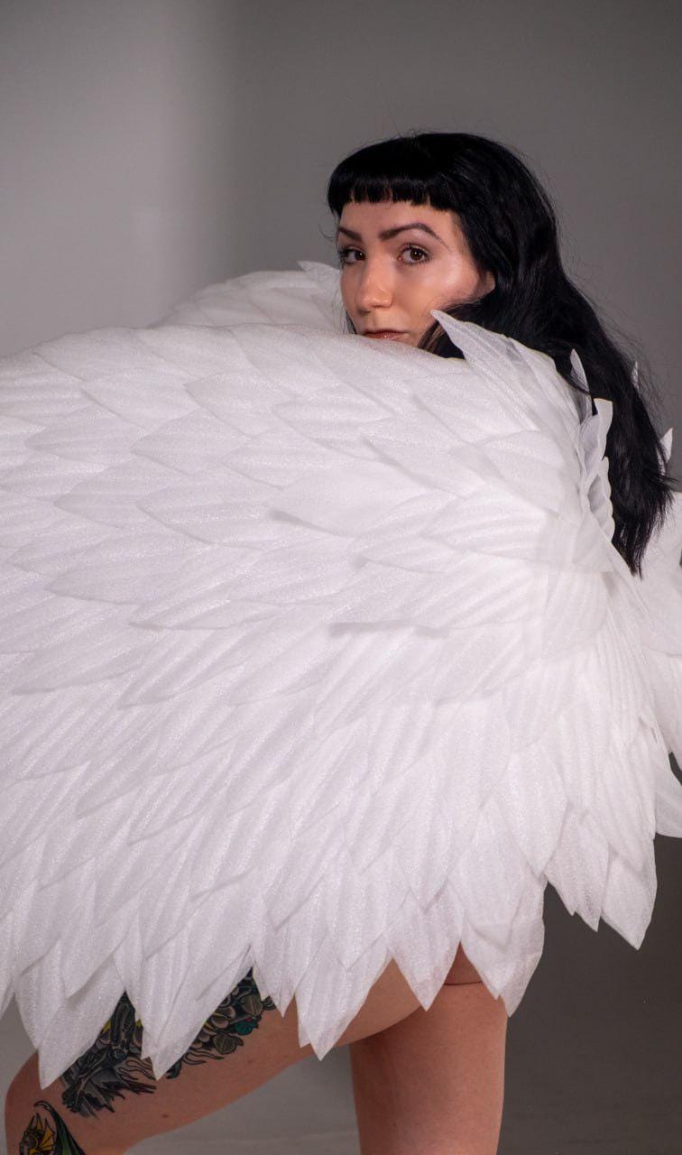 May include: A person wearing large, white, feathery wings. The wings are made of layered, textured material, creating a voluminous effect. The person has dark hair and is looking over their shoulder.