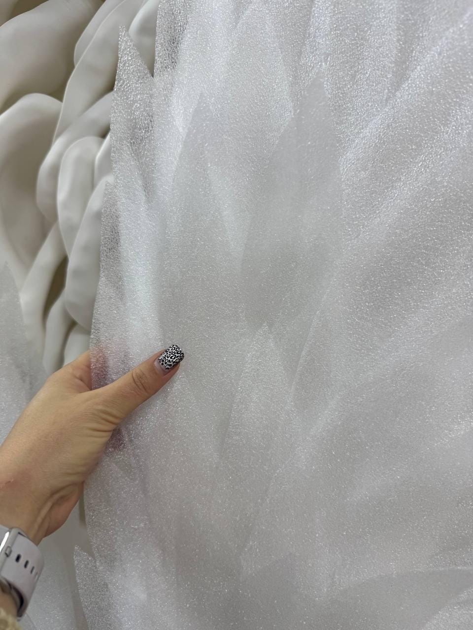 May include: Close-up of white tulle fabric with a textured, semi-transparent appearance. The fabric is being held, revealing its delicate, airy quality. The background shows more of the same fabric, creating a sense of depth and volume. The fabric is ideal for crafting and fashion.