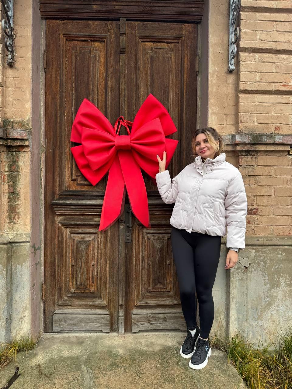 May include: A large, bright red bow is mounted on a dark wooden door. The bow is the focal point, with its voluminous loops and long, flowing tails. The door is part of a building with brick and stone elements.