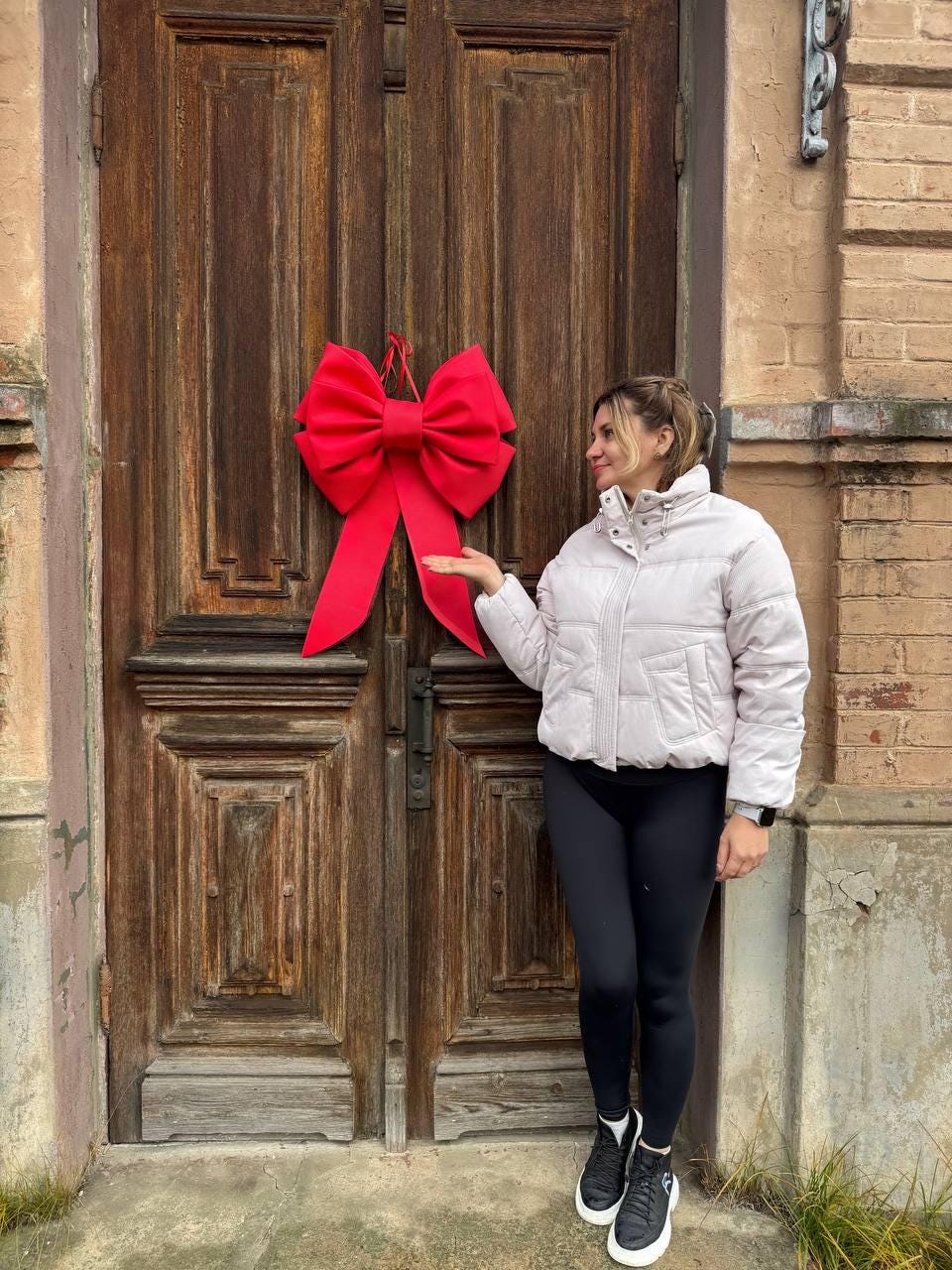 May include: A large, bright red bow is hanging on a weathered wooden door. The bow is the focal point, with long, flowing tails. The door has a rustic, aged appearance, with visible wood grain and panels. The bow adds a festive touch.