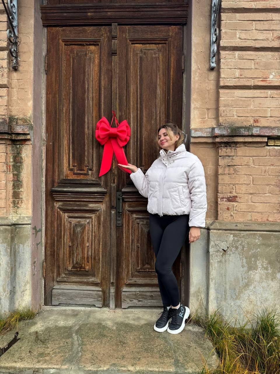 May include: A large, bright red bow is hanging on a weathered wooden door. The door is made of dark brown wood with visible grain and panels. The bow is the focal point, adding a pop of color against the door's rustic texture. The door is set in a brick building.