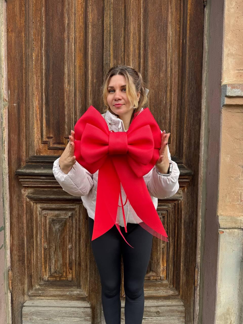 May include: A large, vibrant red bow with long streamers. The bow is held in front of a person, against a backdrop of a wooden door. The bow's color is a bold, festive red.