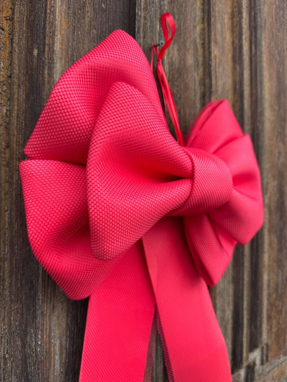 May include: A large, vibrant red bow with a textured surface, hanging against a wooden background. The bow is tied with a long, flowing ribbon, perfect for festive decorations or gift wrapping. The bow is approximately 12 inches wide.